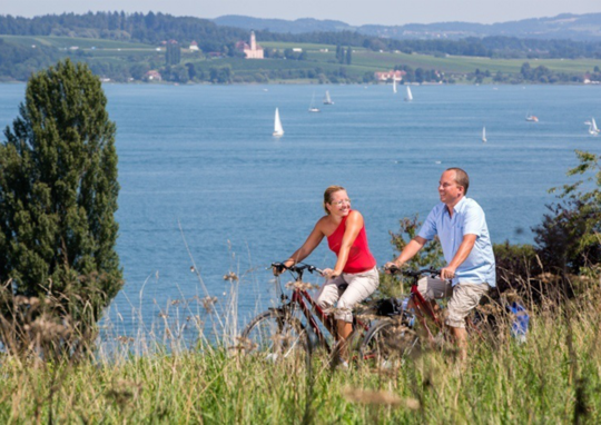 Ein Pärchen fährt auf Fahrrädern, im Hintergrund der Bodensee